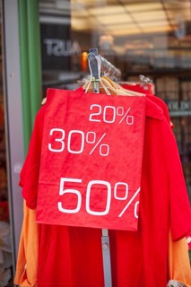 Red sign discount on a shopping bag in front of a clothing rack with t-shirts and shop window,