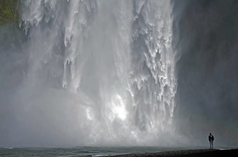 A man stands in front of an impressive waterfall, surrounded by splashing spray, Skogafoss,