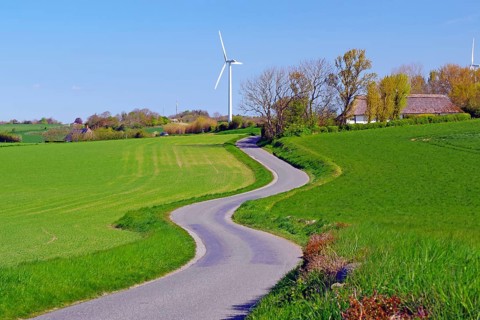 A winding road leads through green fields with wind turbines in the background, cycle tourism,