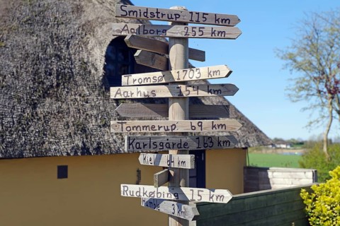 A wooden signpost indicates various distances, next to a house with a thatched roof, Langeland,