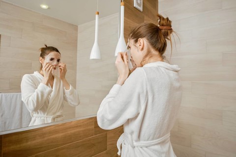 A woman in a white robe is applying skincare mask to her face, standing before a large mirror in a