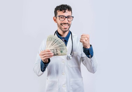 Smiling young doctor holding money isolated. Cheerful doctor holding dollars bills isolated