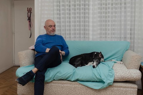 A mature man sits comfortably on the sofa in his home, accompanied by his greyhound. The sofa