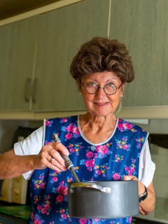 Granny with old smock apron, glasses and wig in the kitchen, holding cooking pot and ladle,