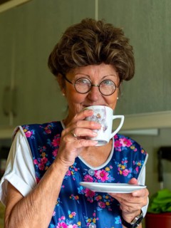 Granny with old smock apron, glasses and wig in the kitchen, holding coffee cup, Germany, Europe