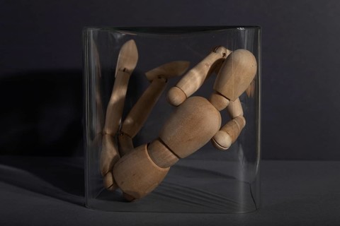Wooden mannequin locked in a glass cube on gray background. captivity, break rules, loneliness,