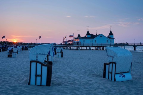Beach chairs on the sandy beach, people strolling on a pier with buildings in the sunset, clear,