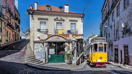 A yellow tram on a cobblestone street passing by a vintage building under a clear blue sky, Alfama,
