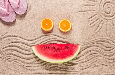 Summer wallpaper, drawing on sand, sea waves and sun, face shape of summer fruits, pink flip-flops,