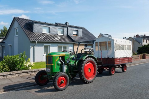 A green tractor with red wheels pulls a white trailer in front of a house under a blue sky, Ilsede,