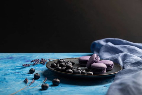 Purple macarons or macaroons cakes with blueberries on ceramic plate on a blue and black background