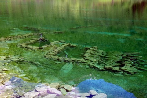 Clear, green water with visible stones and ruins underneath, surrounded by natural landscape,