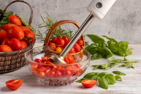 A blender in a glass bowl with sliced tomatoes, surrounded by fresh tomatoes in baskets, basil and