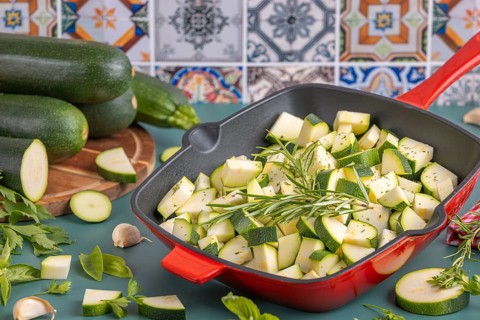 Diced courgettes in a red pan, decorated with fresh herbs in front of colourful tiles and stacked