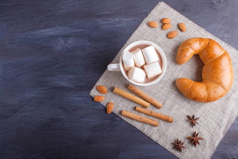 A cup of hot chocolate with marshmallow, croissant and spices on linen napkin and black wooden