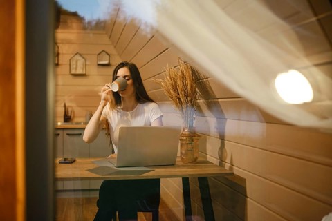 View through the window at a woman sitting at a table and drinking tea in wooden house in the