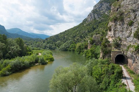 Natural riverbank at the foot of an impressive rock face under a cloudy sky, railway tunnel, Nestos