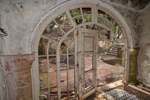 An abandoned building with an arched doorway, broken window panes and broken stone steps, conveying