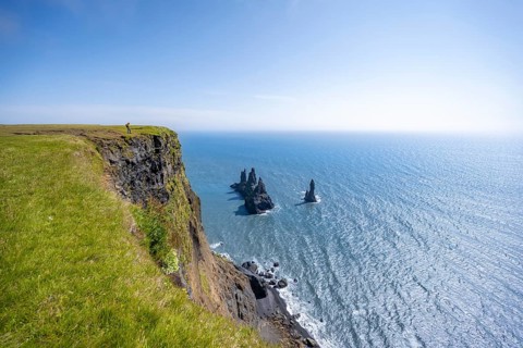 Young man standing on cliff, view from steep cliff to the rocks Reynisdrangar in the water, at
