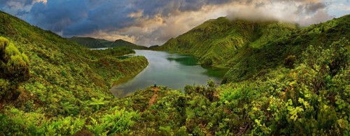 Panoramic view of a lake surrounded by green forested mountains under a dramatic sky, crater lake