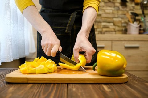 Unrecognizable man in apron slicing bell peppers on wooden cutting board in the kitchen