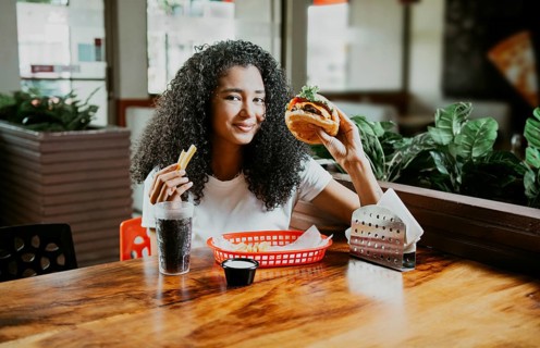Happy girl enjoying a delicious hamburger sitting in a restaurant. Portrait of young afro woman
