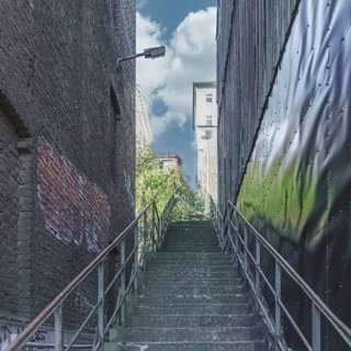 A staircase leads through a narrow alley into bright sunlight, flanked by brick walls and steel