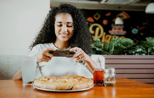 Smiling young woman using phone photographing a pizza in a restaurant. Happy girl photographing a