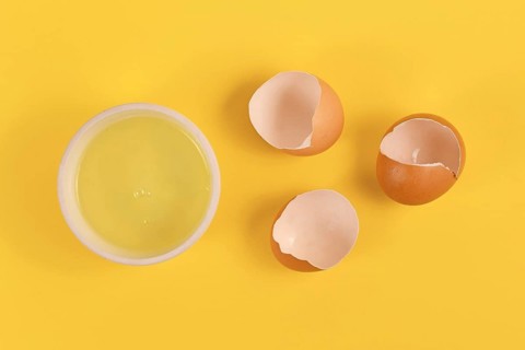 Bowl with egg white next to broken eggshells on yellow background
