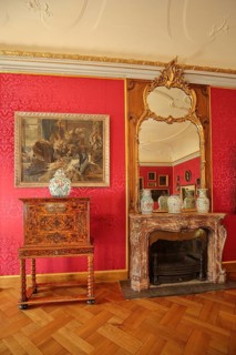 Red drawing room with chest of drawers, painting, wall mirror, parquet floor and fireplace,