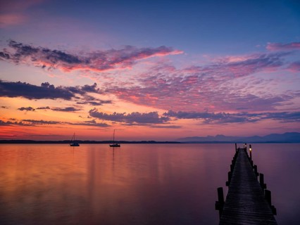 Illuminated clouds over sailing boats and jetty at Chiemsee at dawn, behind the Chiemgau Alps,