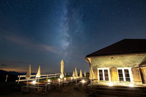 A mountain hut under a clear starry sky with the Milky Way visible and mountains in the background,