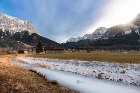 Frozen river on a meadow with dried grass, wooden cabins, a village, and the snow-capped Alps