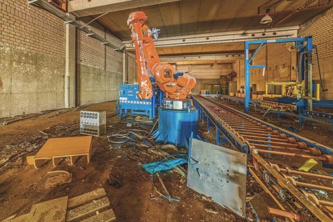 Abandoned factory building with a large orange-coloured robot arm amidst machines and equipment,