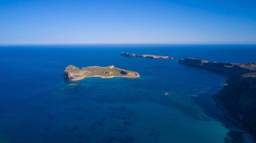 Drone shot, aerial view of an island with green area surrounded by blue sea and steep cliffs,