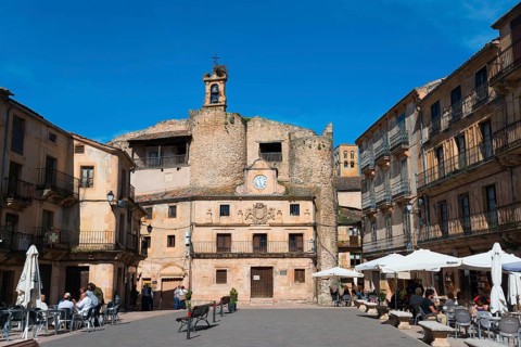 Historic square with stone walls, cafés and parasols, bustling with people and dominated by a clock