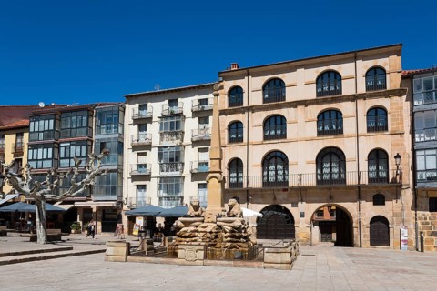 A quiet square with a fountain in the centre, surrounded by historic buildings under a clear blue