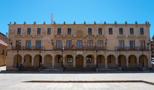 A large historic building with arcades and flags stretching across a spacious square under a blue