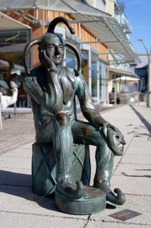 Bronze statue of a pensive jester sitting on a cube and holding a mask next to him, carnival jester