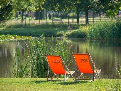 Two orange deckchairs on the shore of a pond in a quiet green park on a sunny day, papenburg,