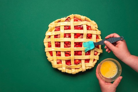 Woman hands washing with egg yolk the pie crust. Top view with strawberries and rhubarb tart.