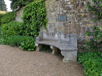 Old curved and ornately designed stone garden seat against old stone wall with green foliage