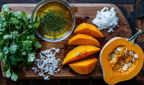 A top-down view of a table with fresh cilantro, pumpkin wedges, minced garlic, and a bowl of herbs