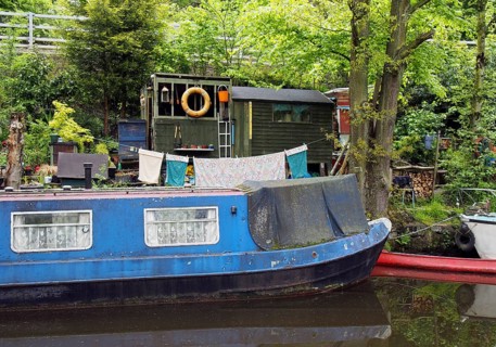 An old scruffy blue narrow boat moored next to wooden sheds with washing on the line on the