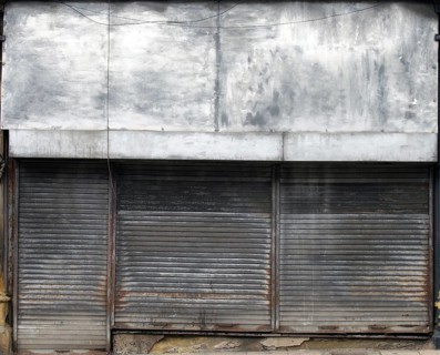 The front of an abandoned store on a street with closed dirty rusting metal shutters over the shop