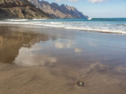 Tidal beach with surrounding mountains and calm sea, waves, clouds reflected in the wet sand,