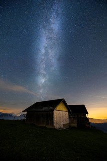 A mountain hut under a clear starry sky with the Milky Way visible and mountains in the background,