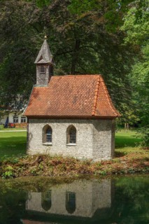 A small stone house with a red tiled roof stands on the edge of a pond, surrounded by dense forest,