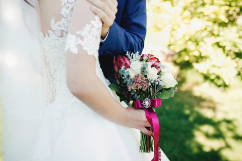 Bride in white dress is holding wedding bouquet. Groom hugs bride. Blurred background.