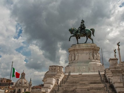 An imposing equestrian statue on a pedestal, flanked by an Italian flag and steps under a cloudy